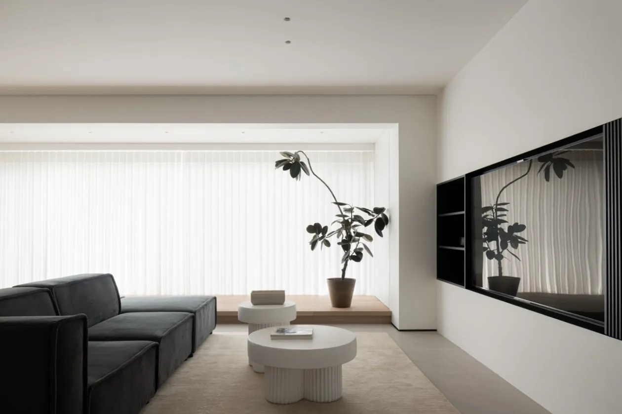 A modern minimalist living room with a black sectional sofa, white coffee tables, a large potted plant, and floor-to-ceiling 