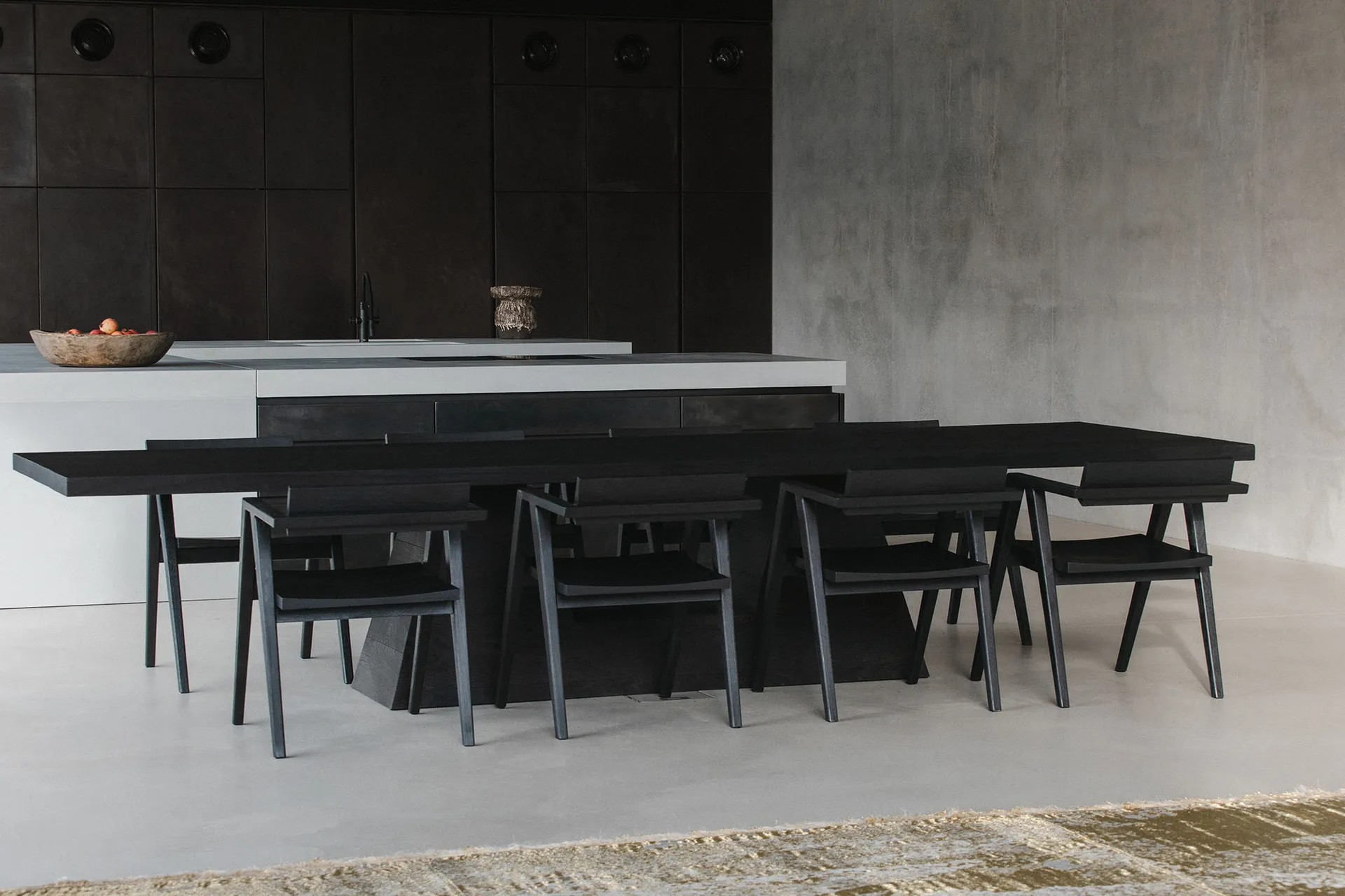 Minimalist kitchen with black furniture, concrete walls, and wood accents. Includes a dining table and chairs.