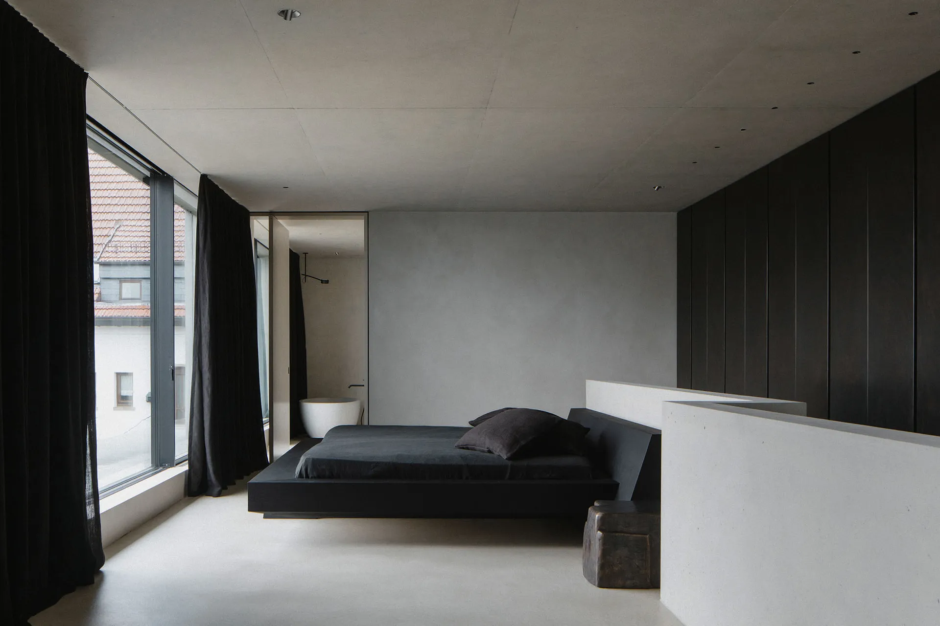 A modern, minimalist bedroom with a concrete floor and walls, featuring a low-profile bed and large windows.