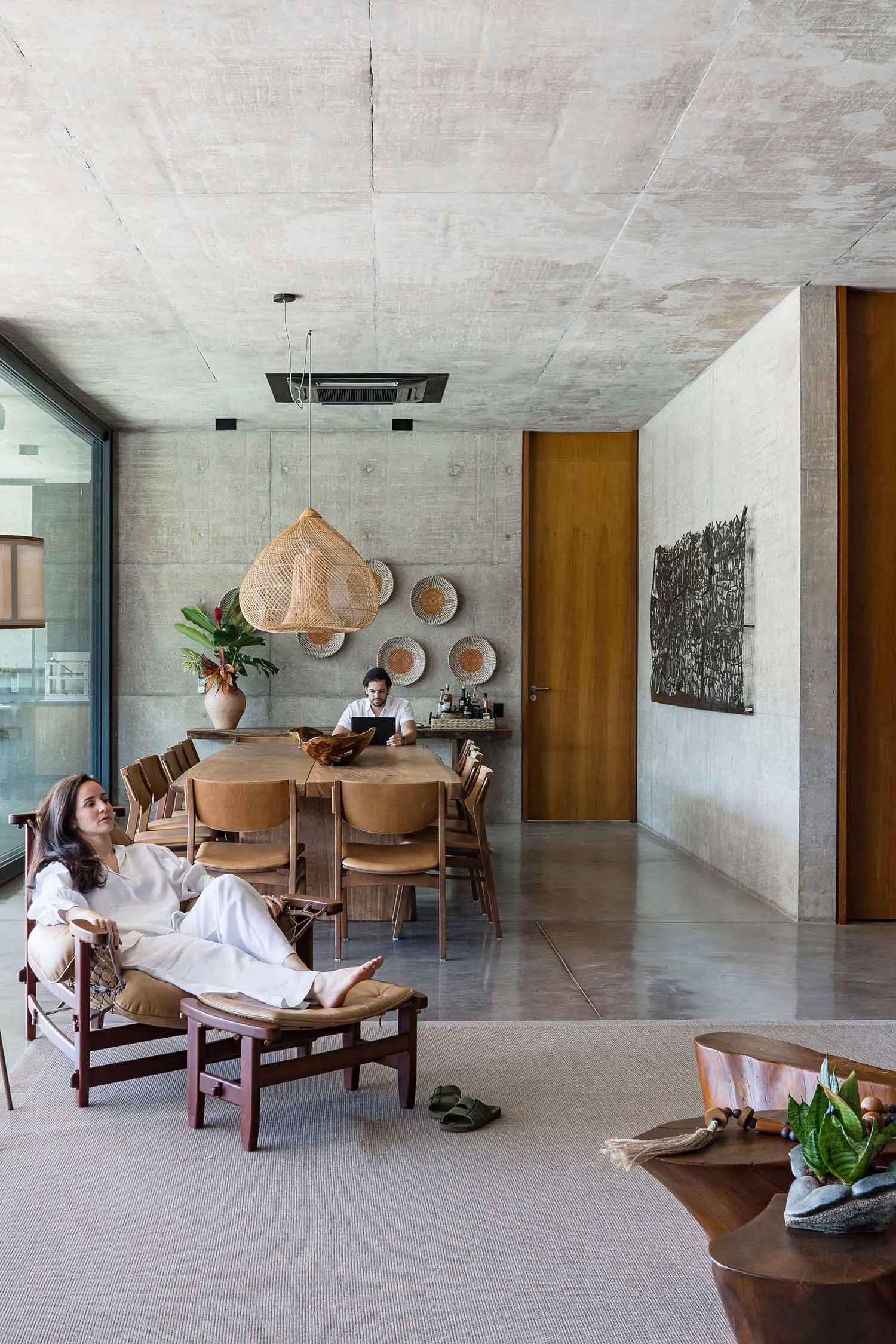 A modern living room with concrete walls, wooden seating, and a large wicker pendant light hanging above. A person is relaxin