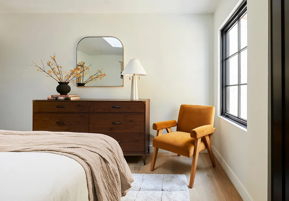 A minimalist bedroom with a wooden dresser, orange chair, and white bedding.