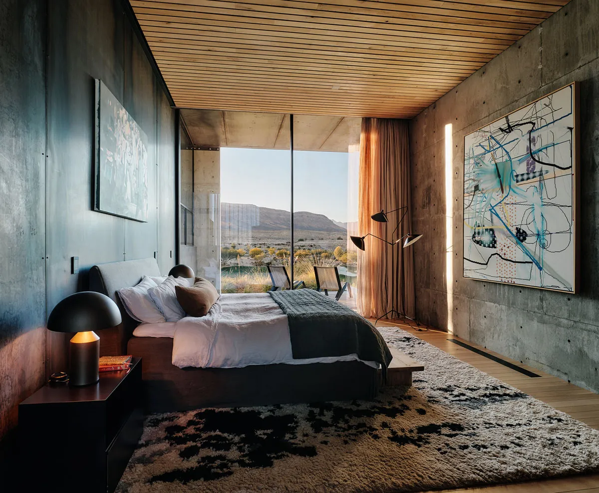 Modern bedroom with concrete walls, wooden flooring, and large glass windows overlooking a natural landscape.