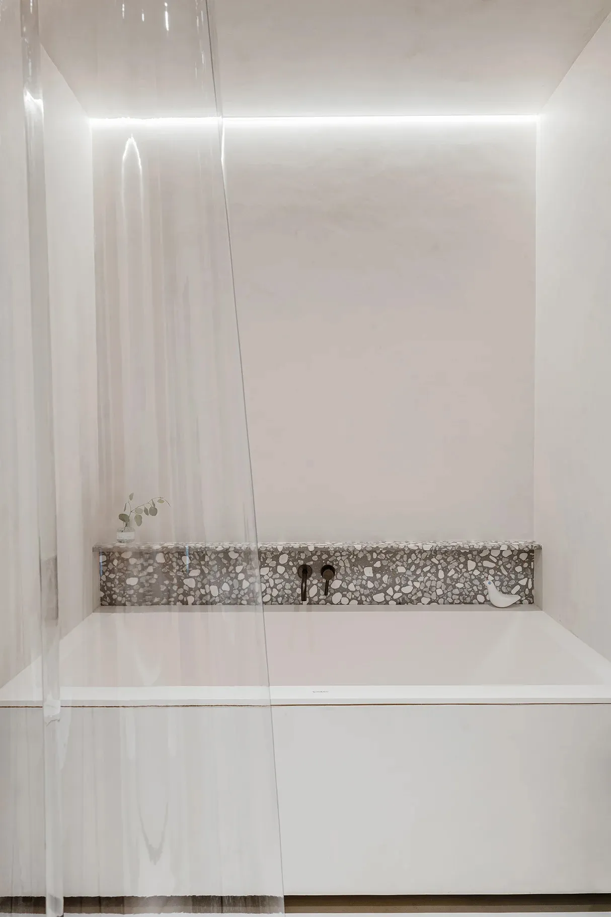 Modern minimalist bathroom featuring a white bathtub with a textured terrazzo backsplash and illuminated lighting.