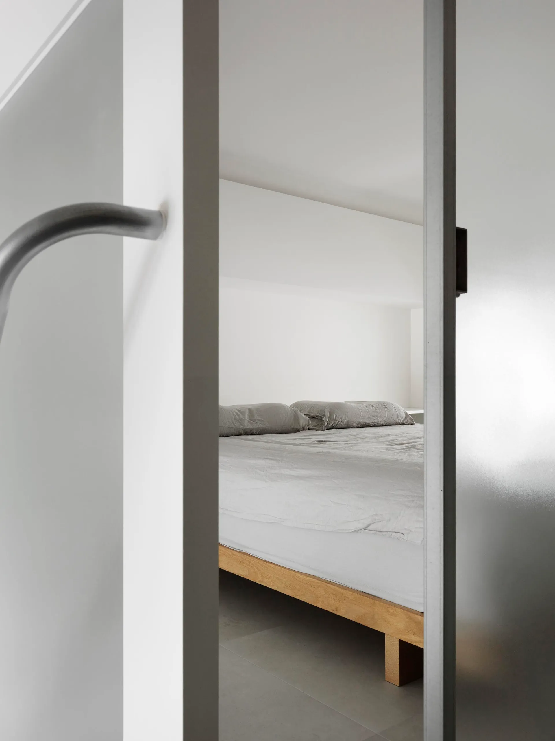A minimalist bedroom featuring a wooden bed frame and grey bedding on a white bed sheet.