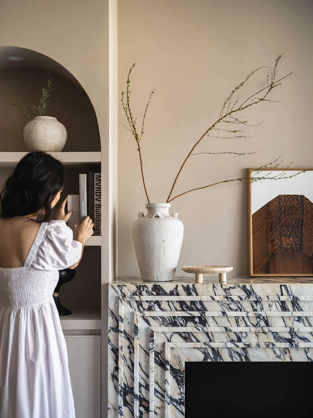 A woman dressed in a white dress is arranging books in a modern living room with a marble fireplace and a vase with branches.