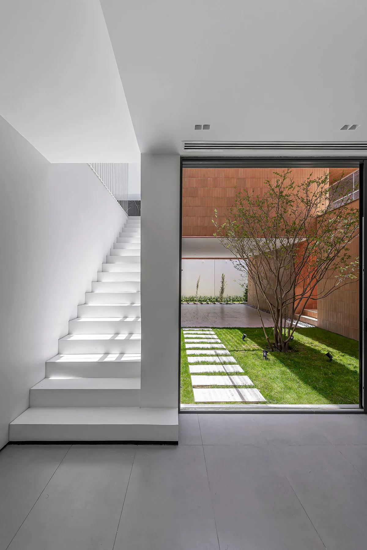 A minimalist interior featuring a white staircase and a glass door leading to a garden with a tree and stone pathways.