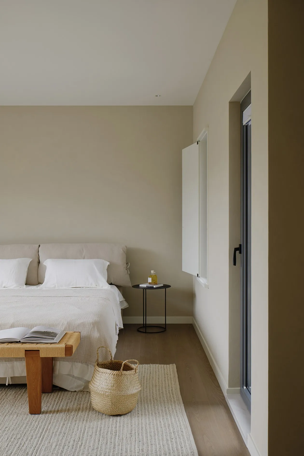 A minimalist bedroom with a neutral color palette, featuring a bed, side table with a bottle, and woven basket on a light-col
