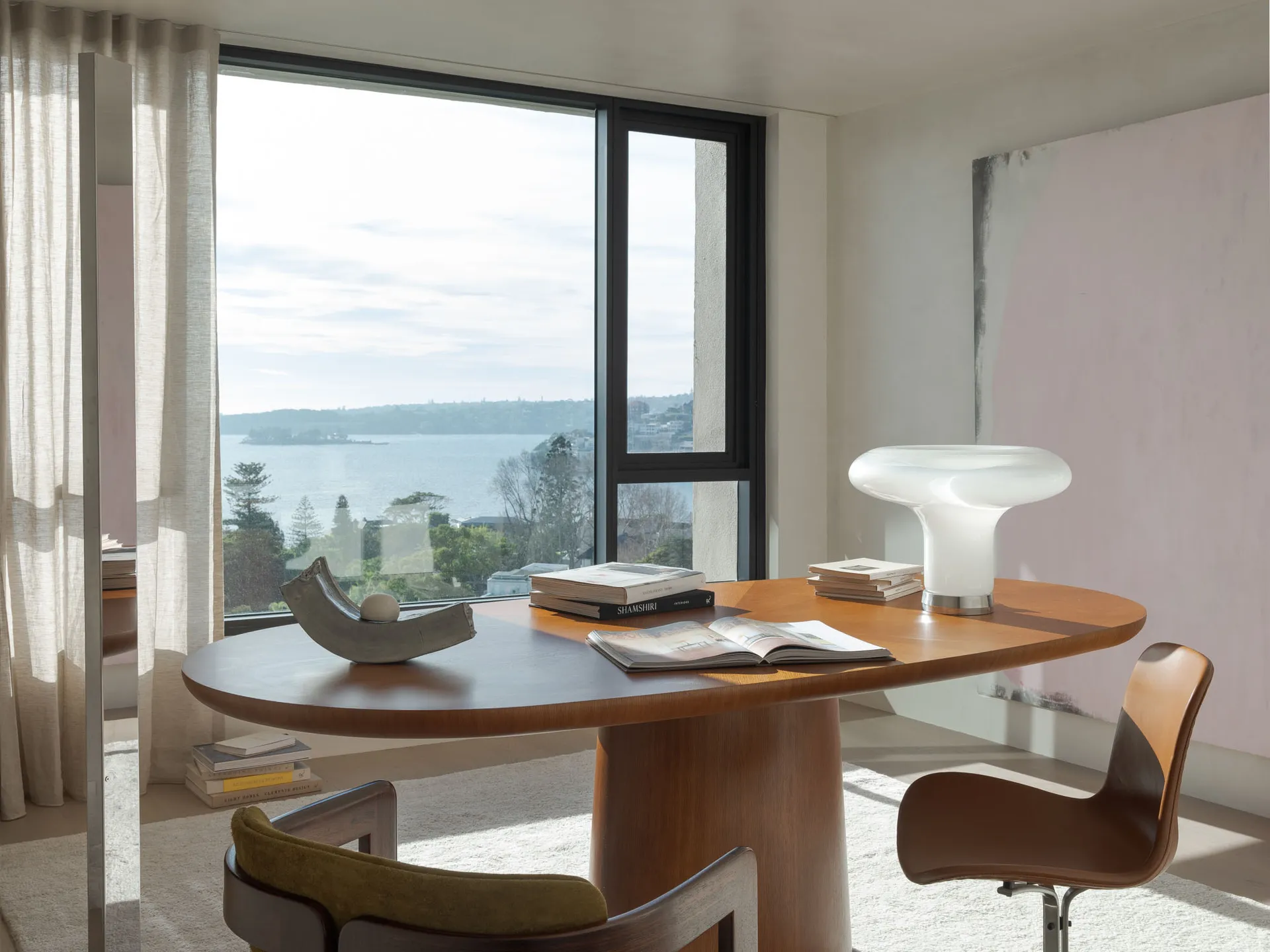 A minimalist home office featuring a wooden table, a white modern lamp, books, and a view of the water through large windows.