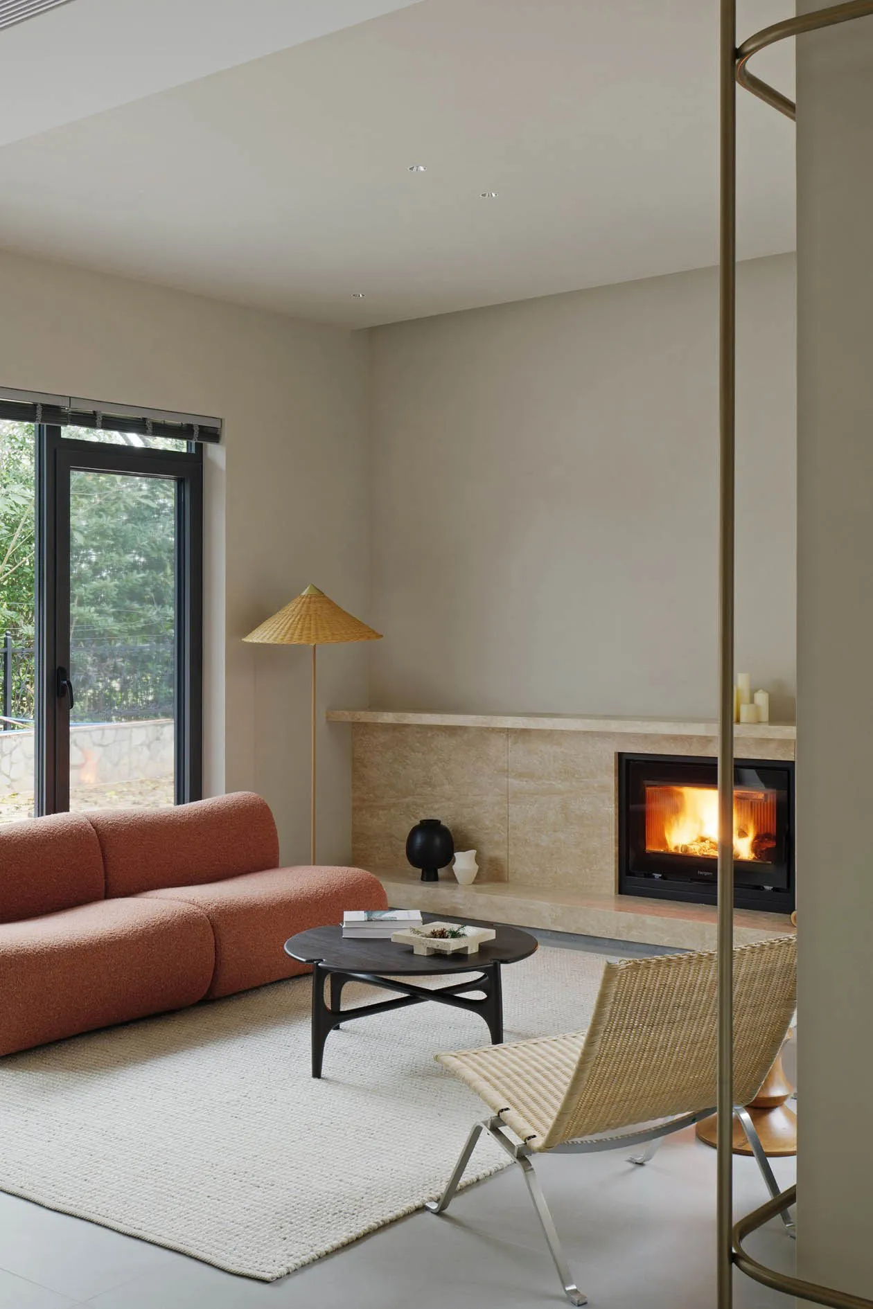 A modern minimalist living room with a fireplace, wicker armchair, and orange sofa in front of a large window with a view of