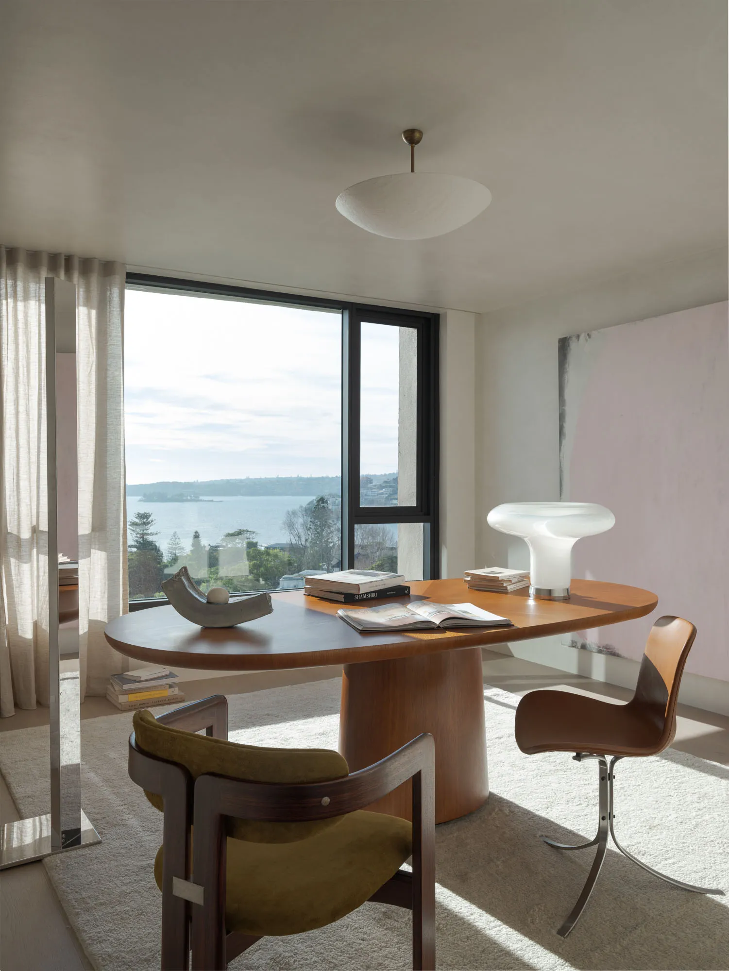 A modern residential office space featuring a wood desk and chairs, with books and a lamp on the desk, and a large window ove
