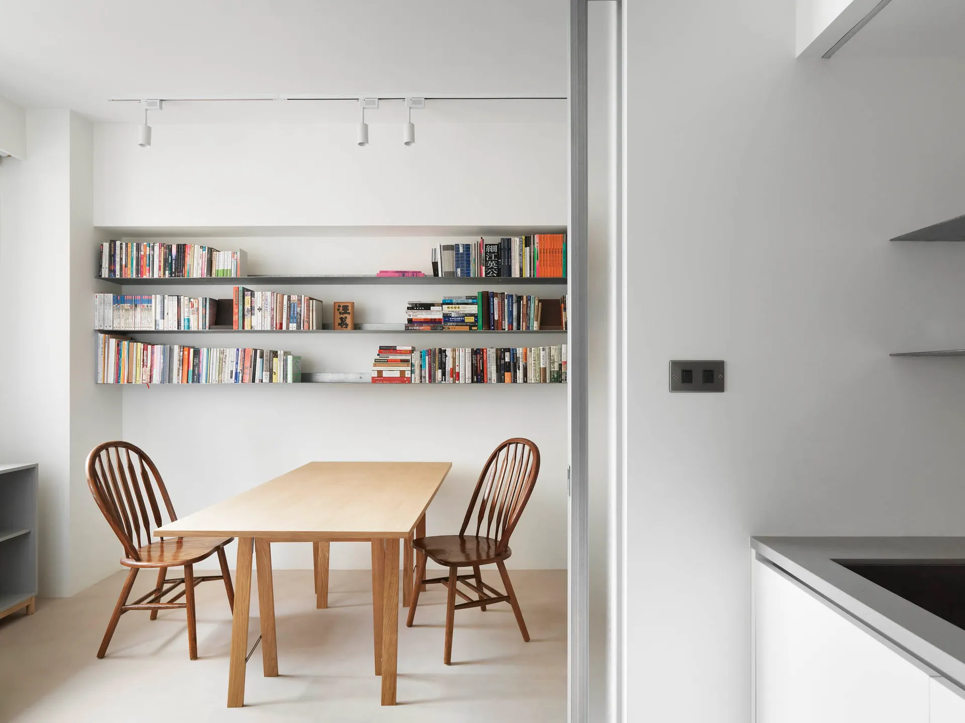 Minimalist dining room featuring a wooden table, wooden chairs, and a metal bookshelf filled with books.