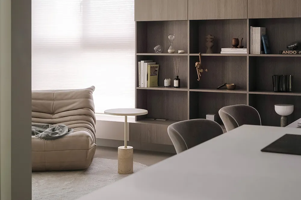 A minimalist living room featuring a beige leather sofa, a white round side table, and a wooden shelving unit with books and