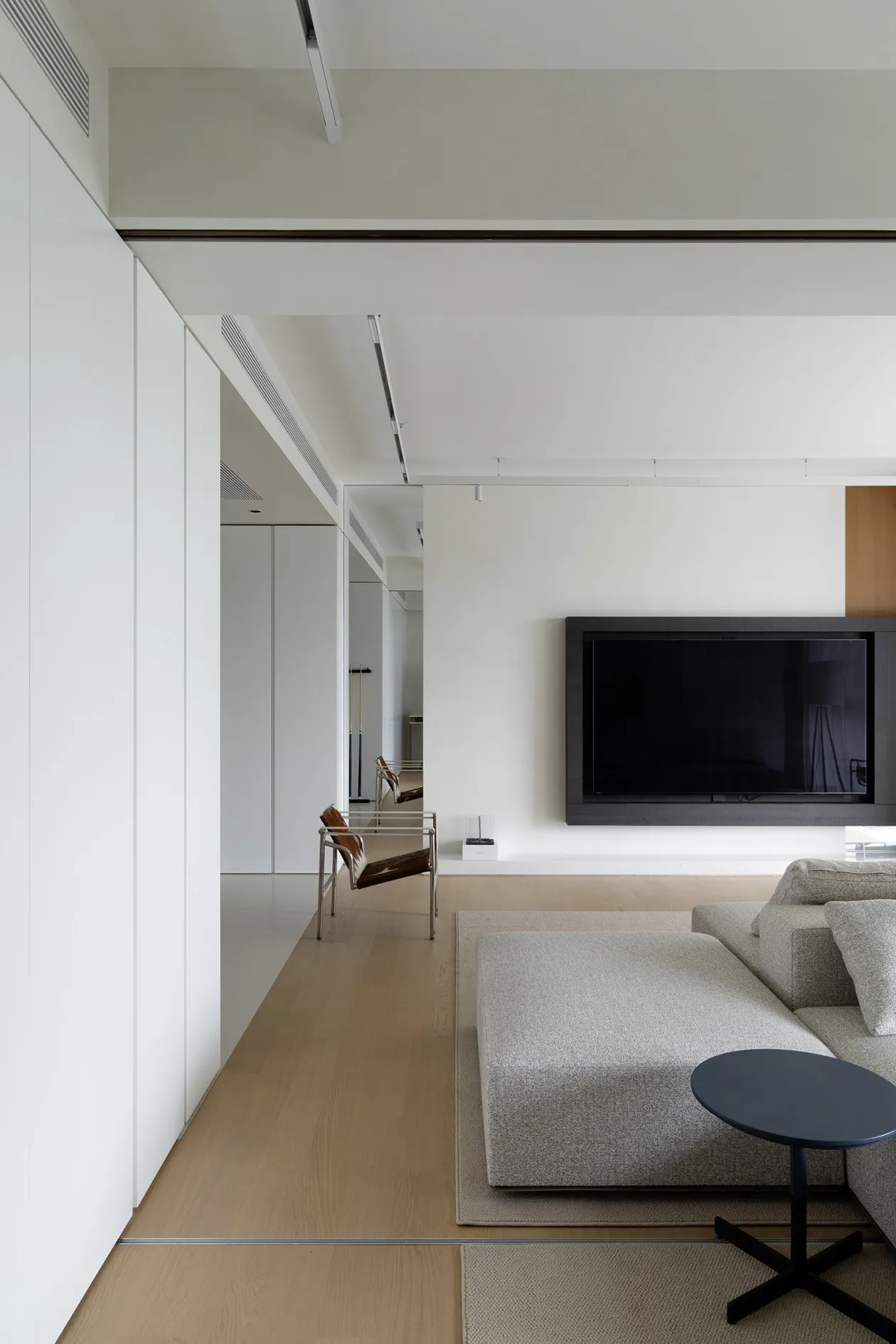 A modern, minimalist living room with white walls, wooden flooring, a leather ottoman, a metal side table, and a large televi