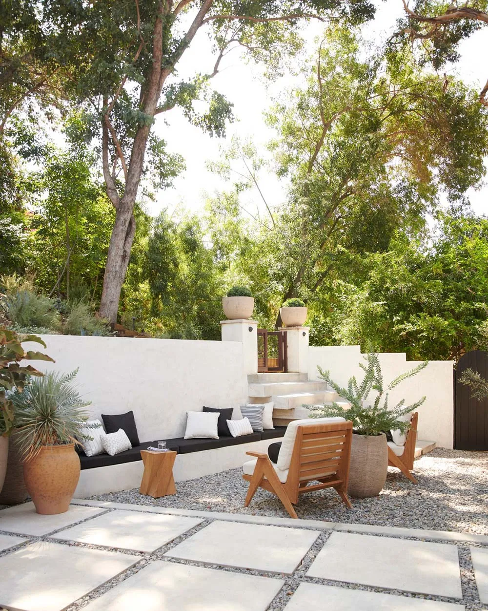 Outdoor patio with modern seating, plants, and a gravel pathway surrounded by greenery and trees.
