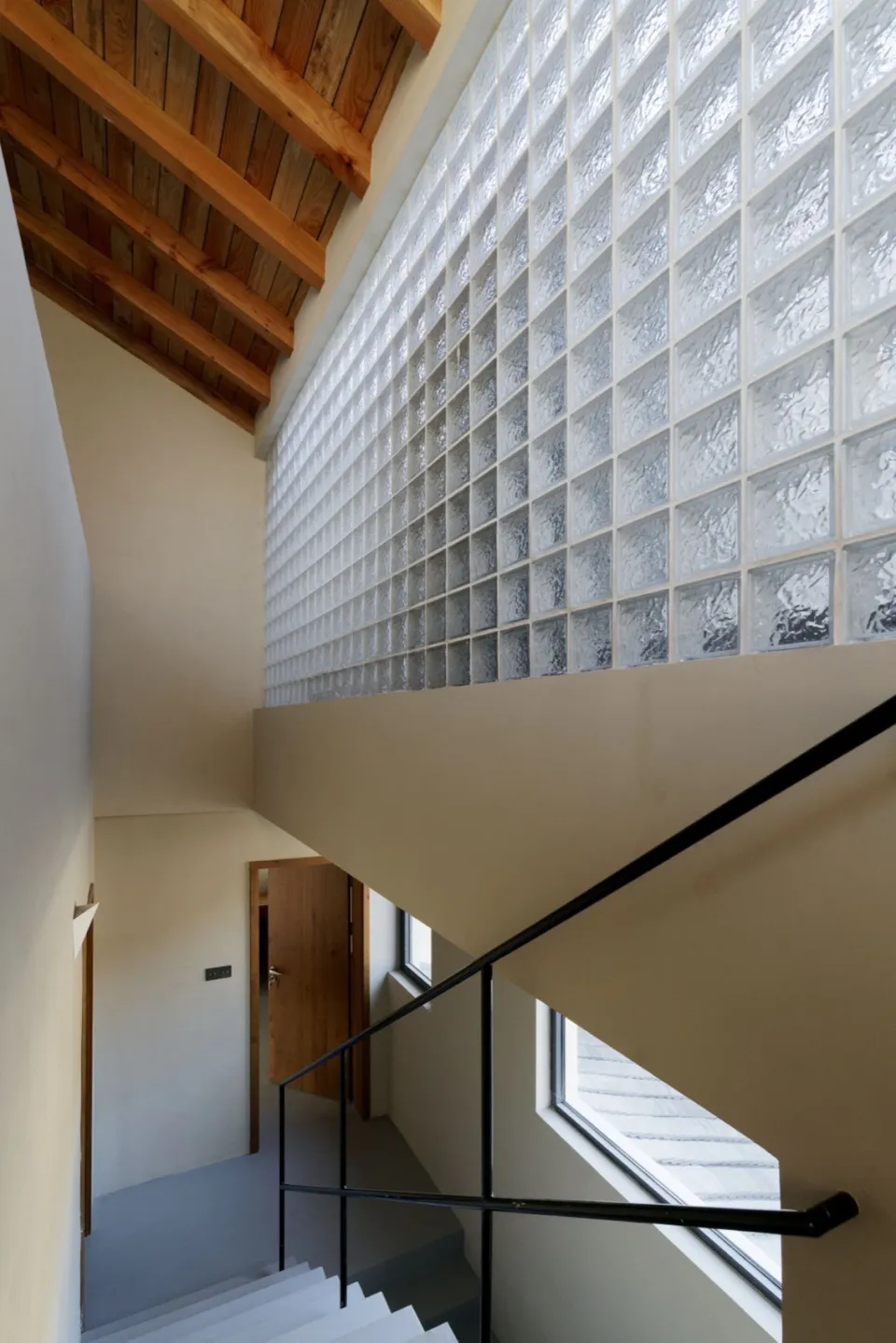 Industrial design staircase with transparent glass blocks on the wall, featuring a wooden ceiling beam and black metal railin