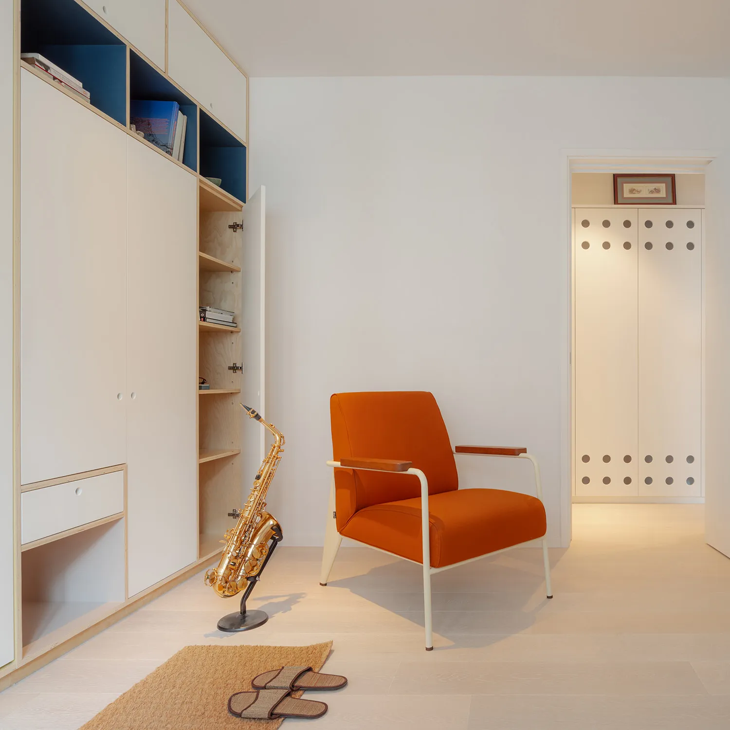 A minimalist living room with an orange upholstered chair, wooden floor, and white cabinetry.