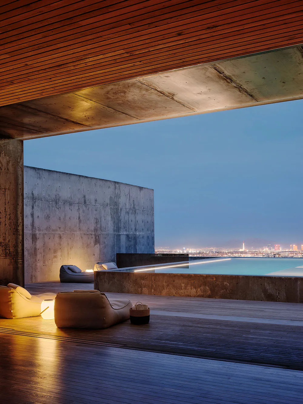 A modern pool deck with concrete walls and floors, featuring a lounge area with beanbag chairs and a view of a cityscape at d