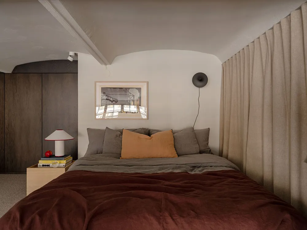 A modern bedroom with mixed materials including a bed with a duvet, pillows, and a headboard. The room features a desk lamp a
