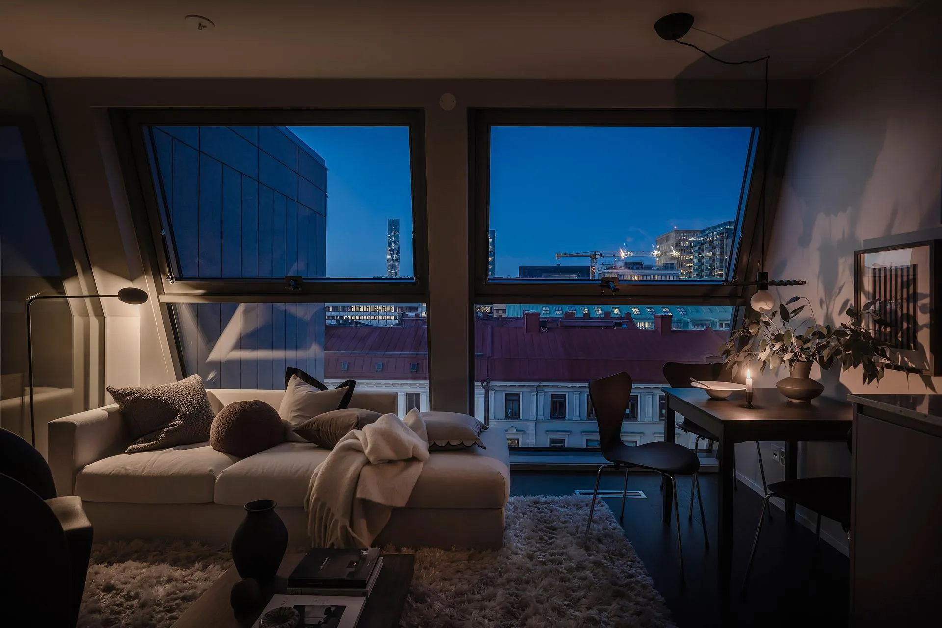 A cozy modern living room with a city view at dusk, featuring soft furnishings and natural light.