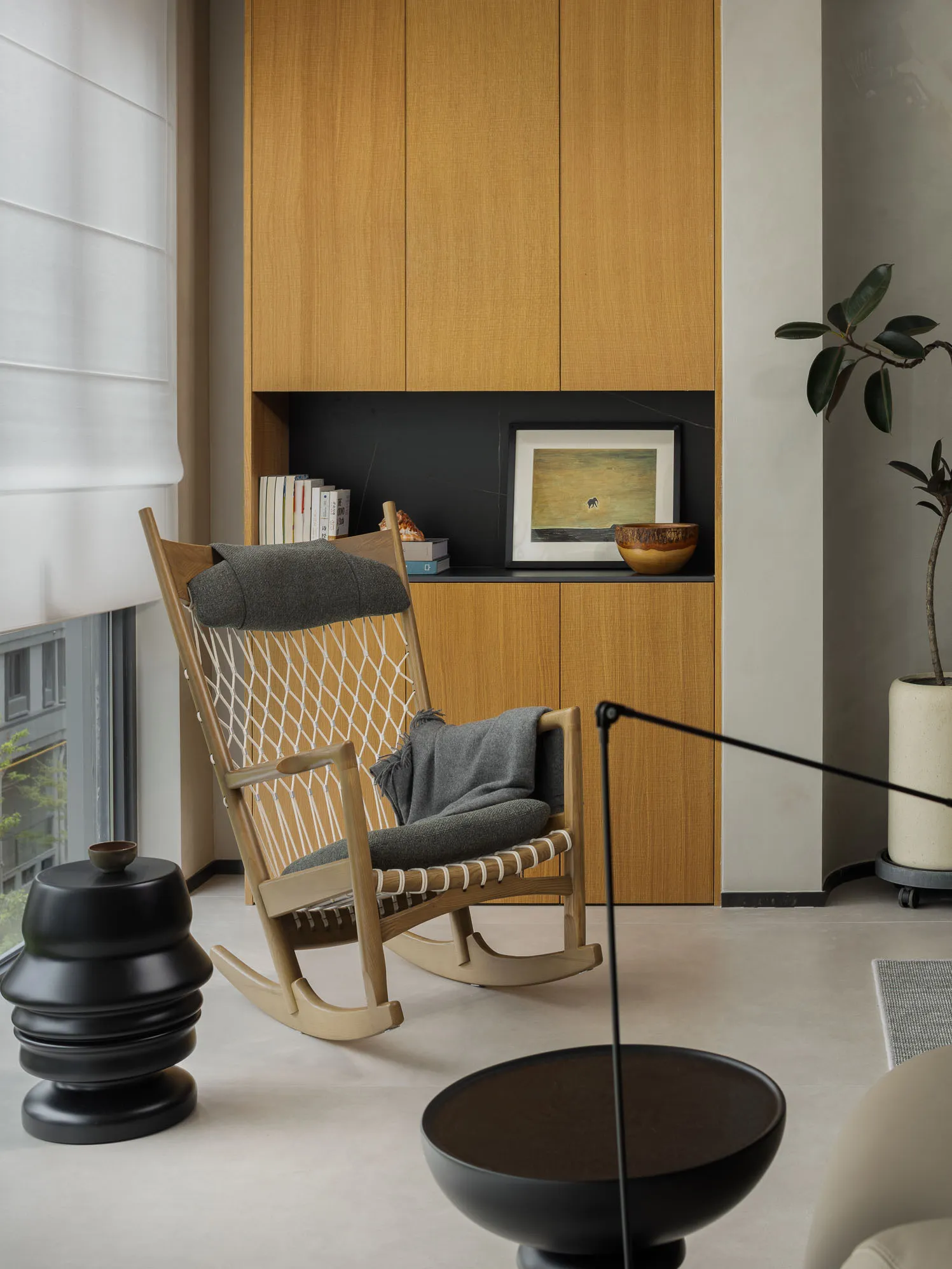 A minimalist living room featuring a wooden rocking chair with a net back, a grey cushion, and a grey blanket.
