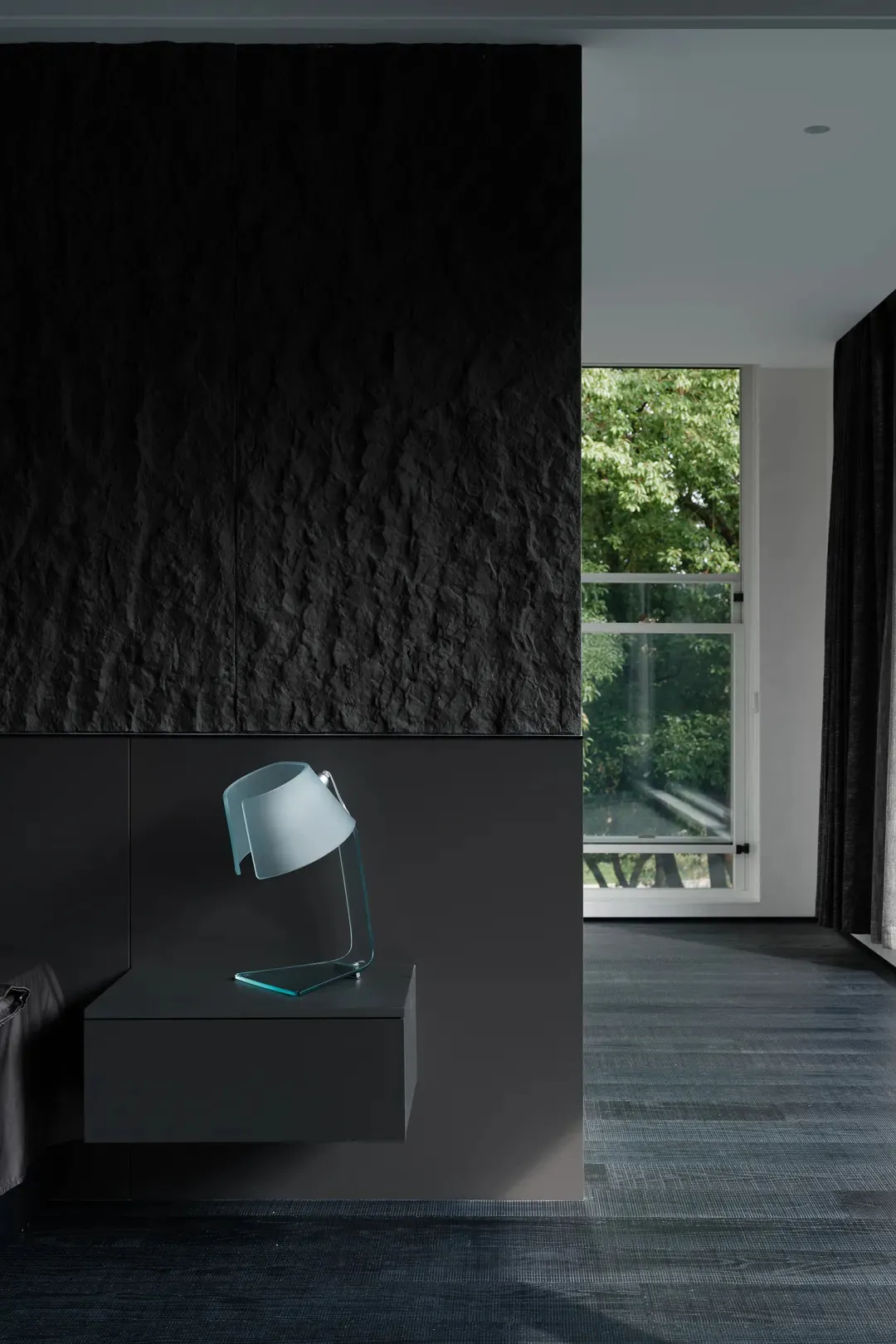 A modern bedroom featuring a glass lamp on a floating nightstand with a textured black wall in the background