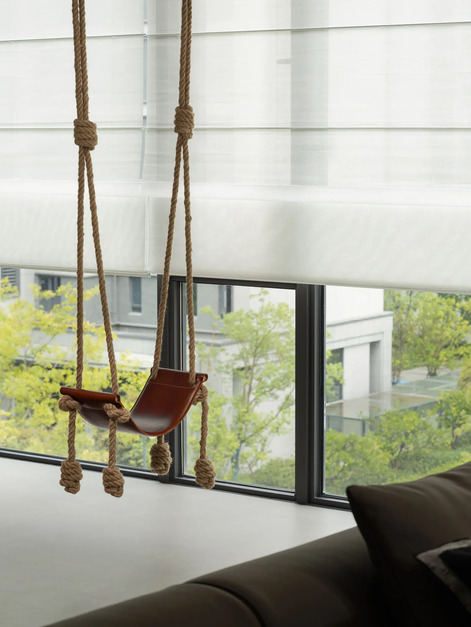 A minimalist living room featuring a hanging leather swing suspended by ropes in front of a large window with white blinds.