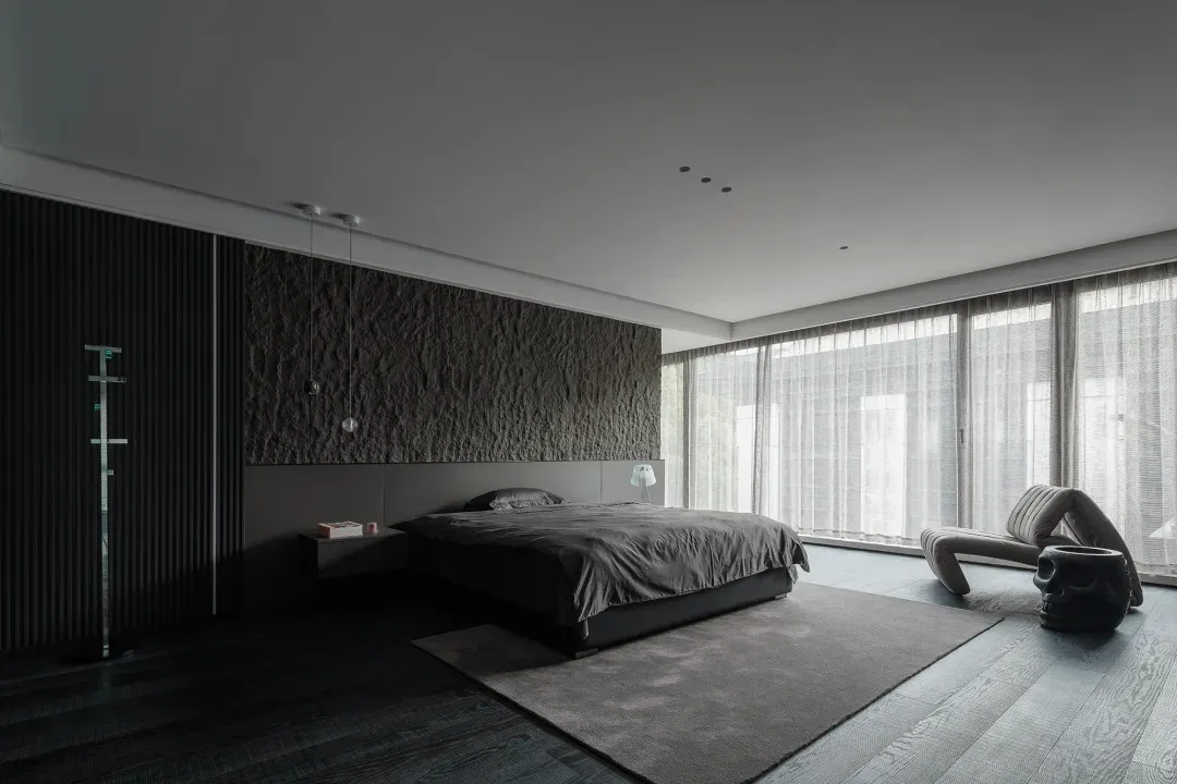 A minimalist bedroom featuring dark wood floors, stone accent wall, and glass walls with a bed and wooden furniture.