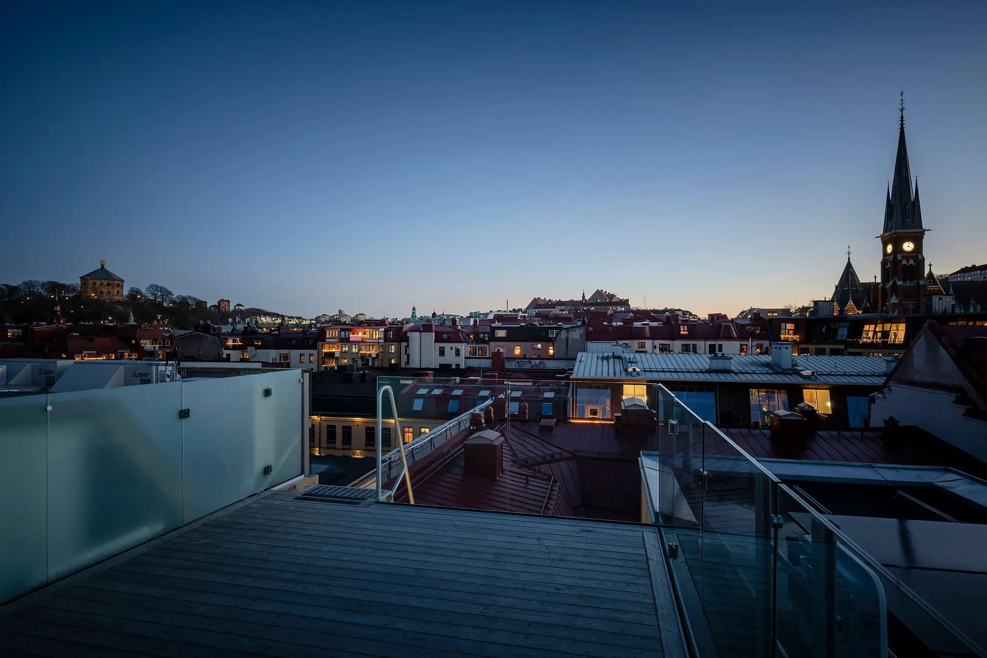 A rooftop view of a cityscape at dusk, featuring illuminated windows and a steeple in the background.