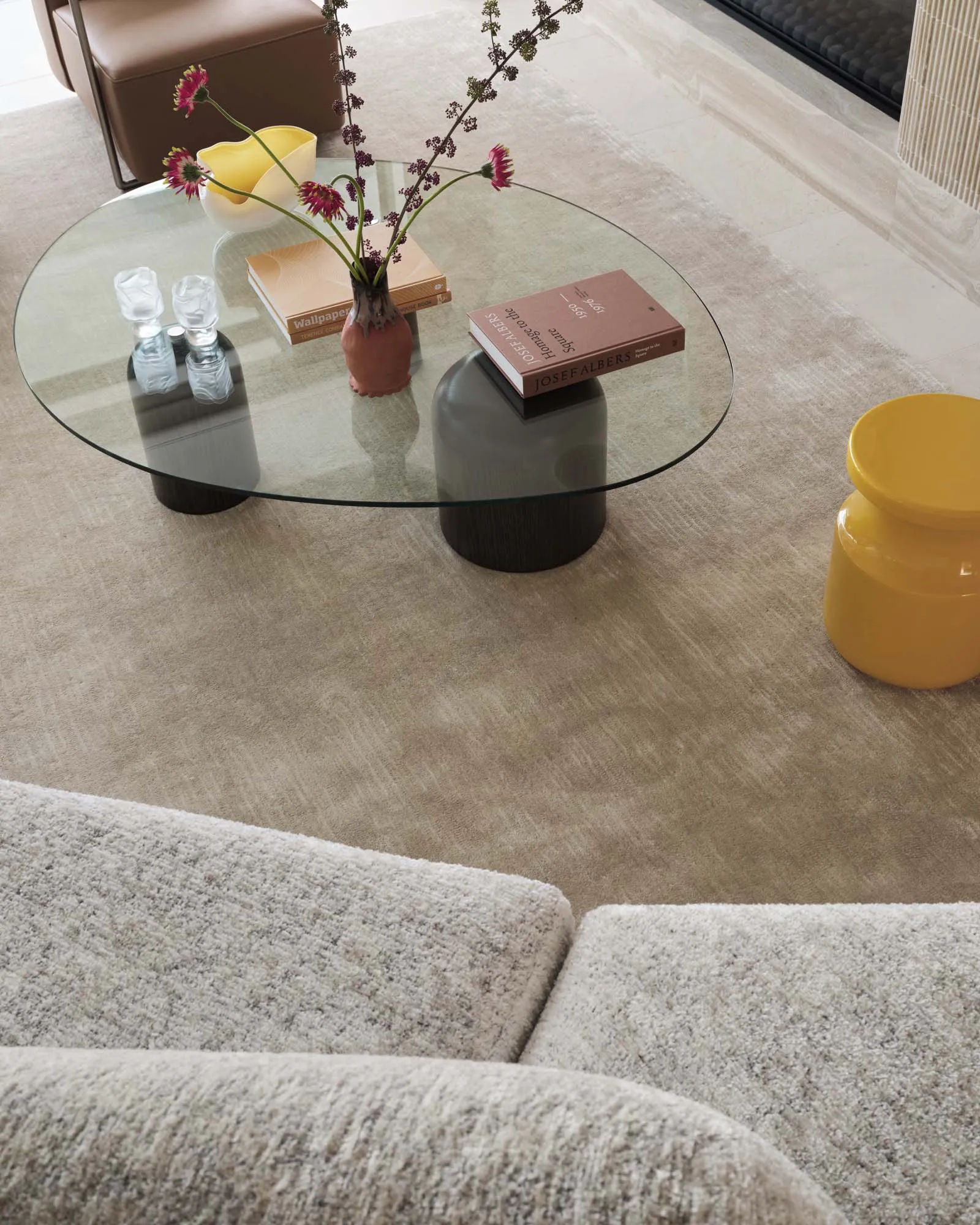 A modern living room featuring a glass coffee table with a vase of flowers and books, placed on a carpet floor with a yellow