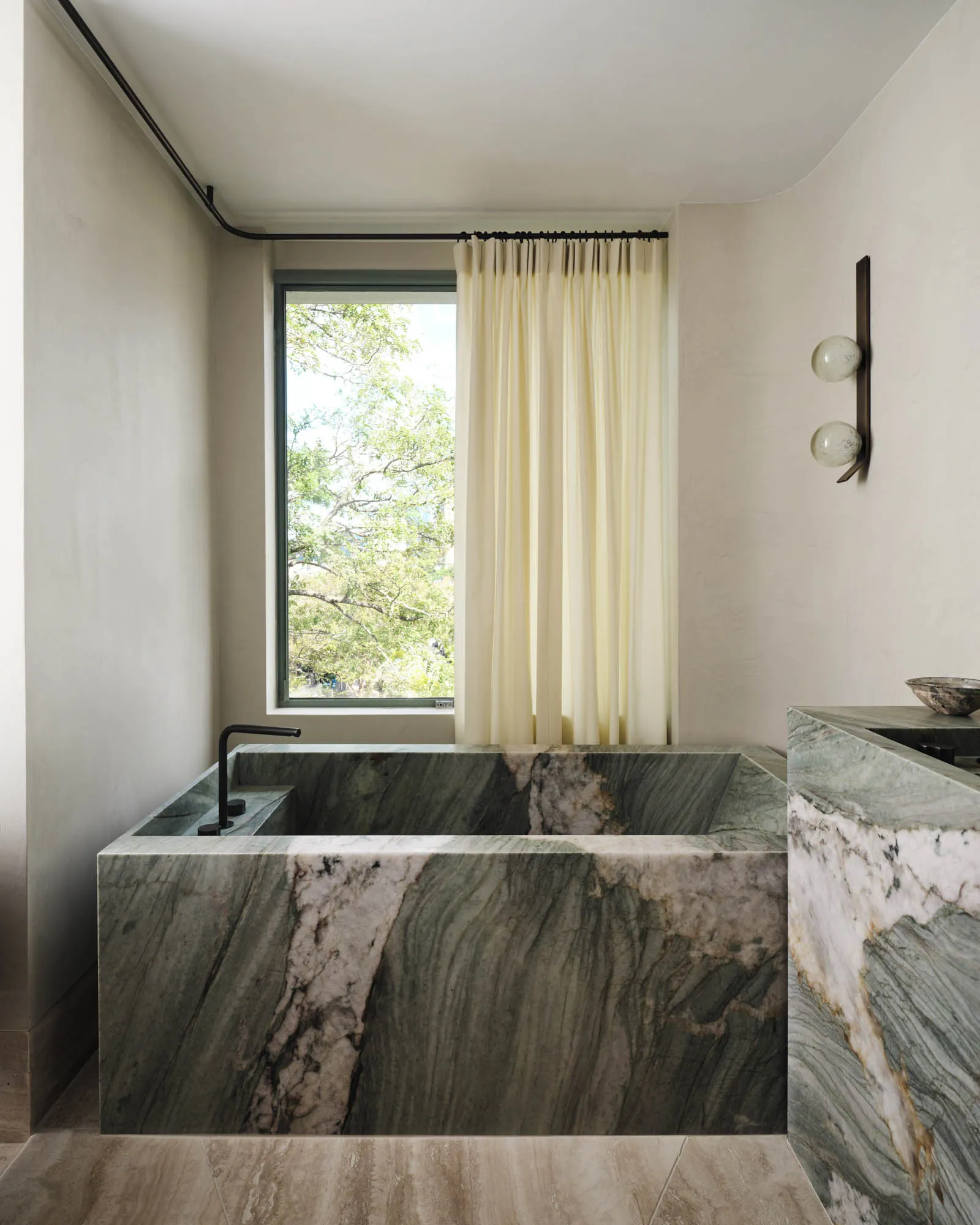 Modern marble bathroom with green and white marble bathtub and floor-to-ceiling curtains by a window.