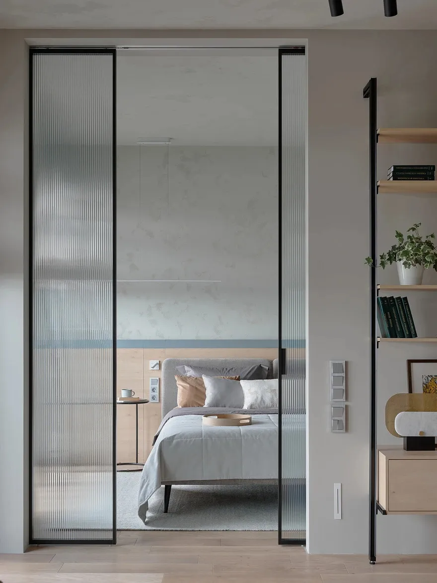 A modern bedroom featuring a clear glass wall, wooden floor, white bed with grey bedding, and a floating shelf with books and