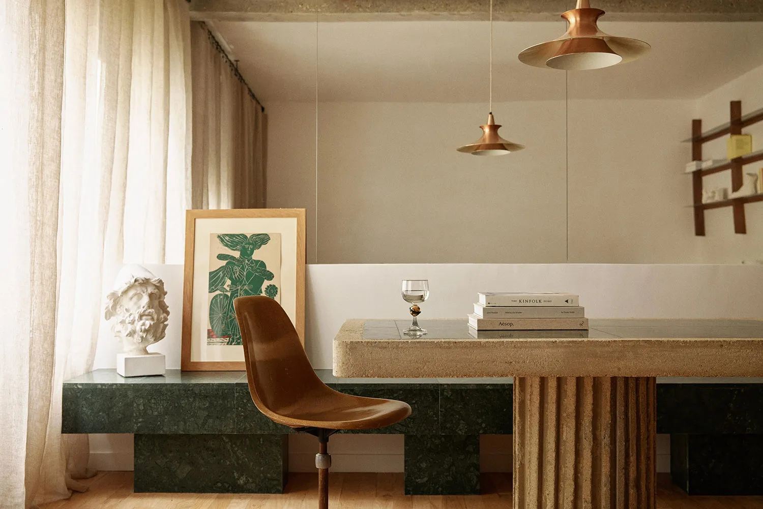 A modern industrial office featuring a concrete and marble desk with a wooden chair, framed artwork, and a glass drink contai