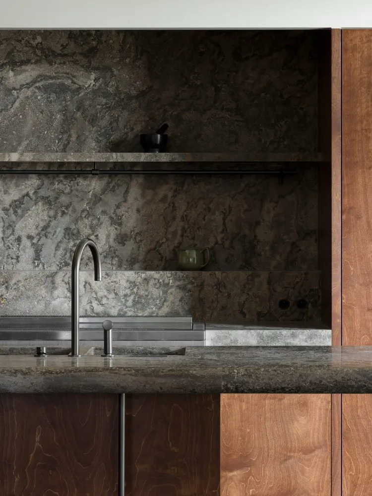 A modern kitchen interior featuring a granite countertop and backsplash, with a metal faucet and wooden cabinets.