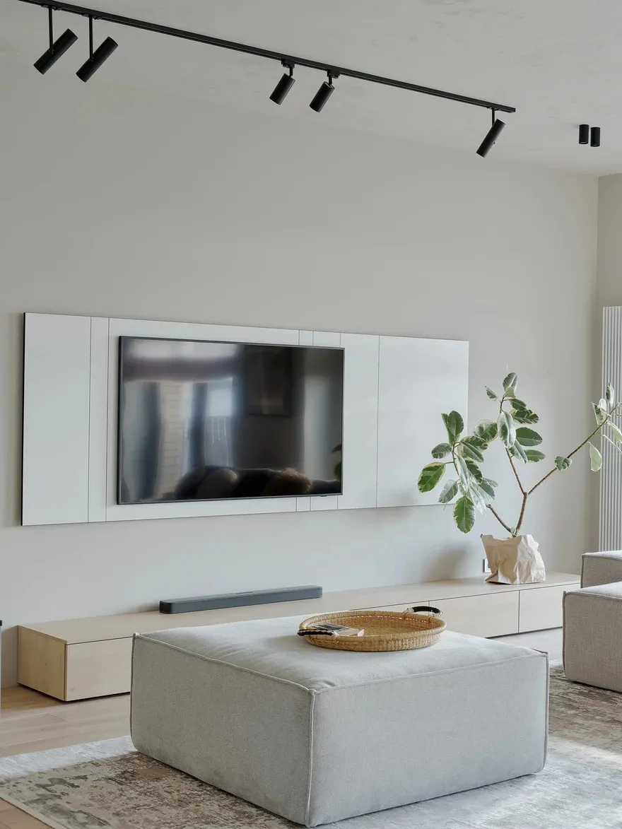A modern living room featuring a light wood floor, minimalist furniture, and a sleek flat-screen TV mounted on the wall.