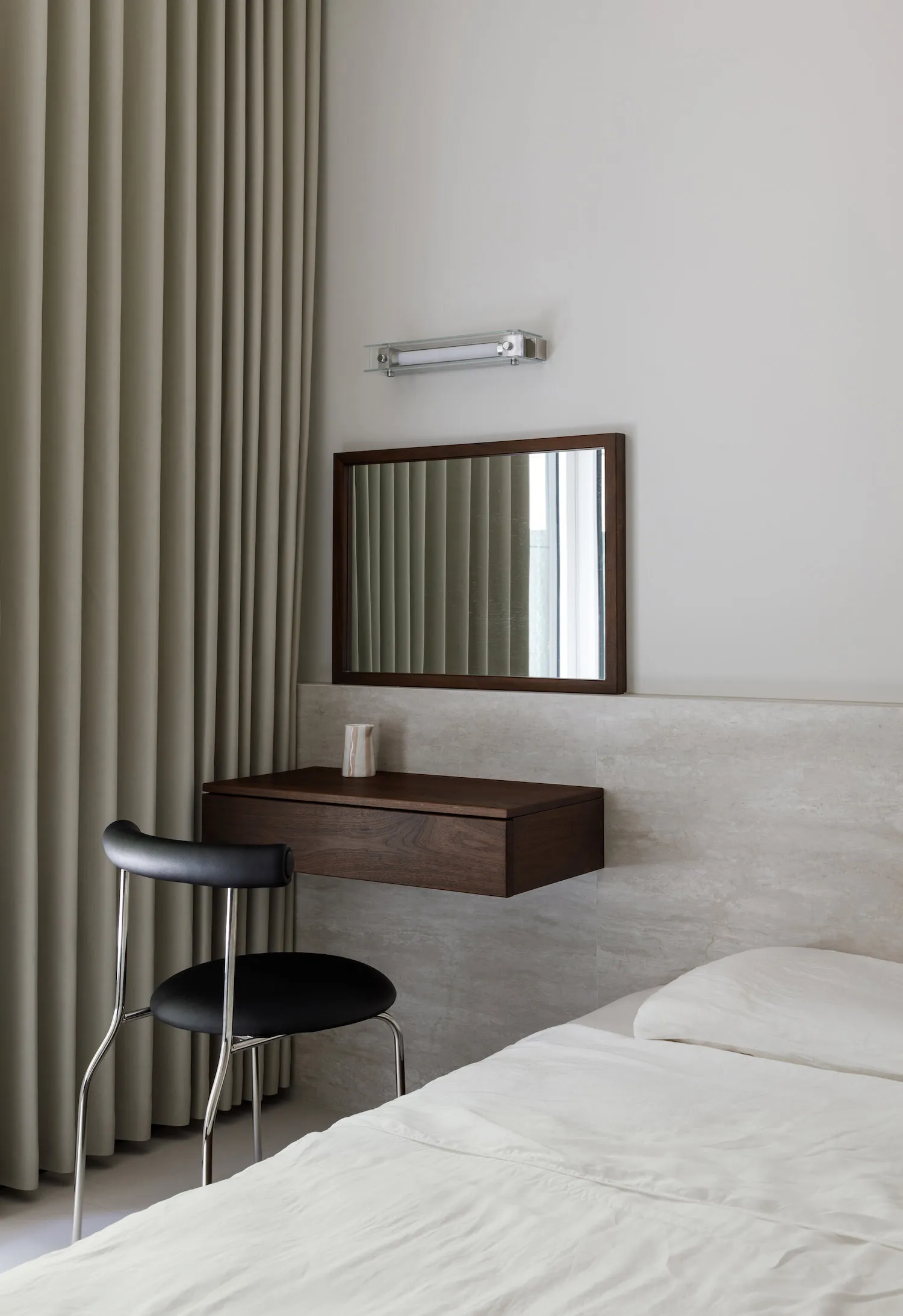 A minimalist bedroom featuring a floating desk with a dark wooden finish, a black chair, and beige curtains.