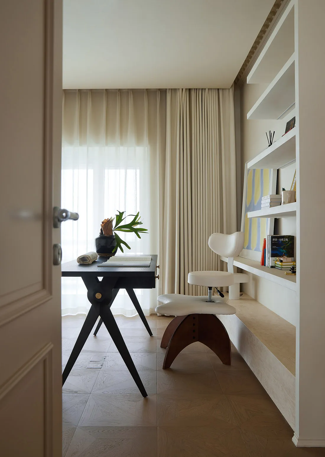 A modern home office space featuring a wooden table and a white desk chair, with a built-in bookshelf and a large window with