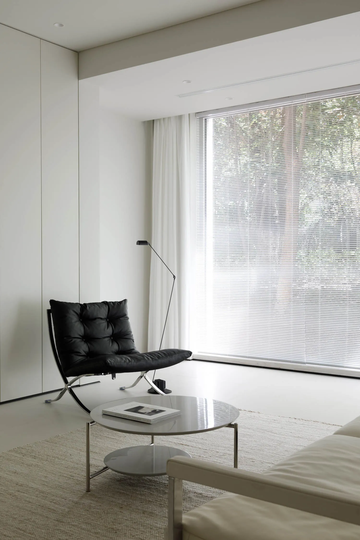 Modern minimalist living room with a black leather chair, white coffee table, and large window with blinds.