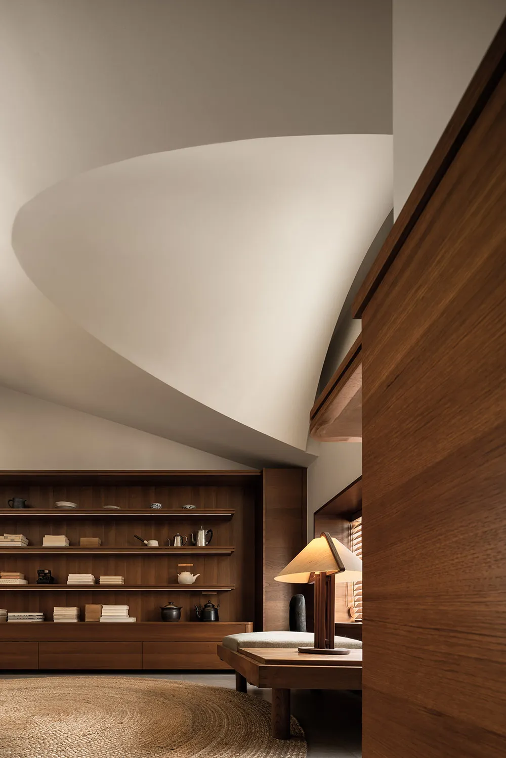 Modern living room with wooden shelves displaying books, vases, and teapots, illuminated by a wooden lamp with a dome-shaped