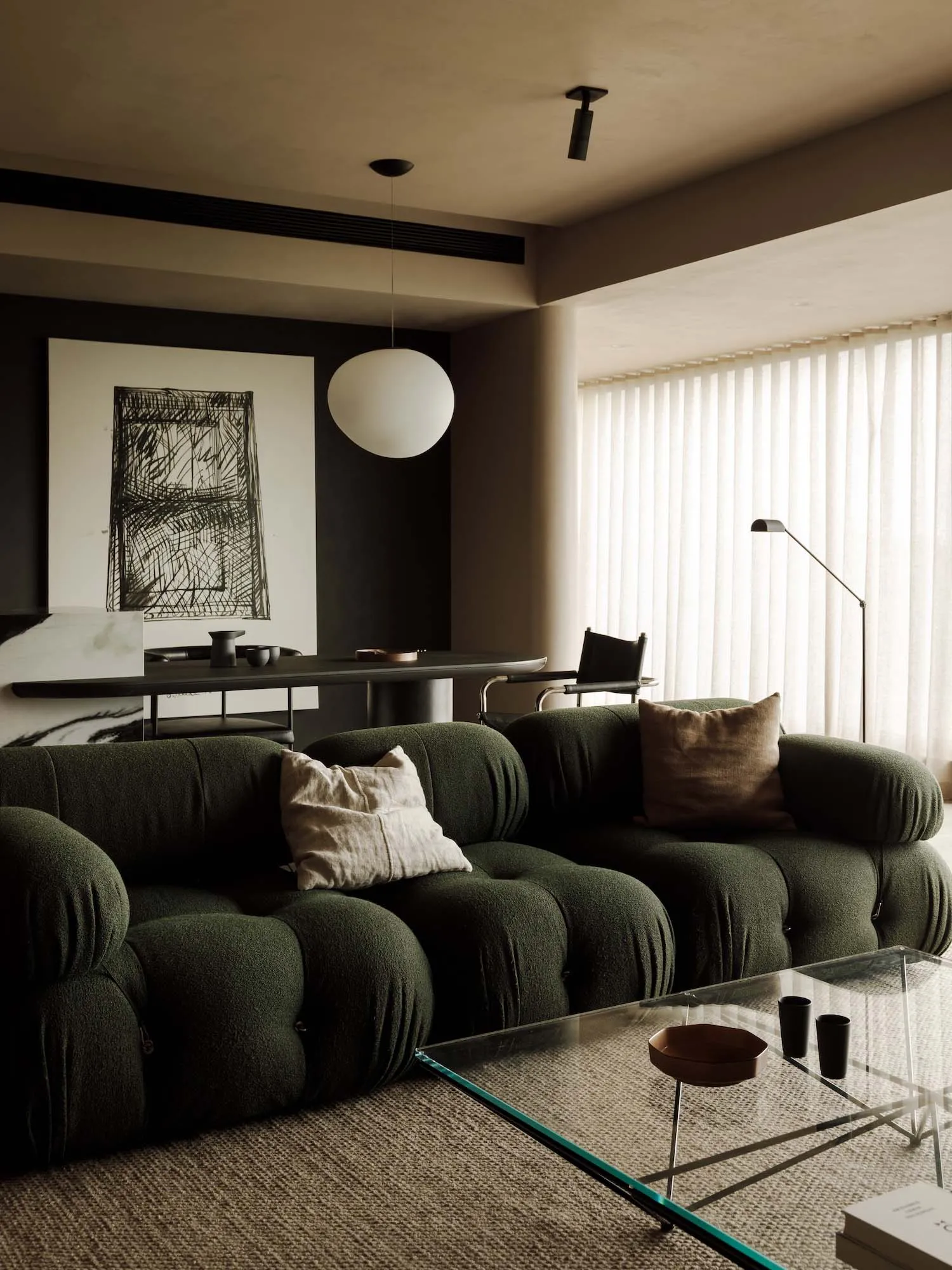 A modern, dark living room with a green fabric sofa, a glass coffee table, and a large window with white curtains.
