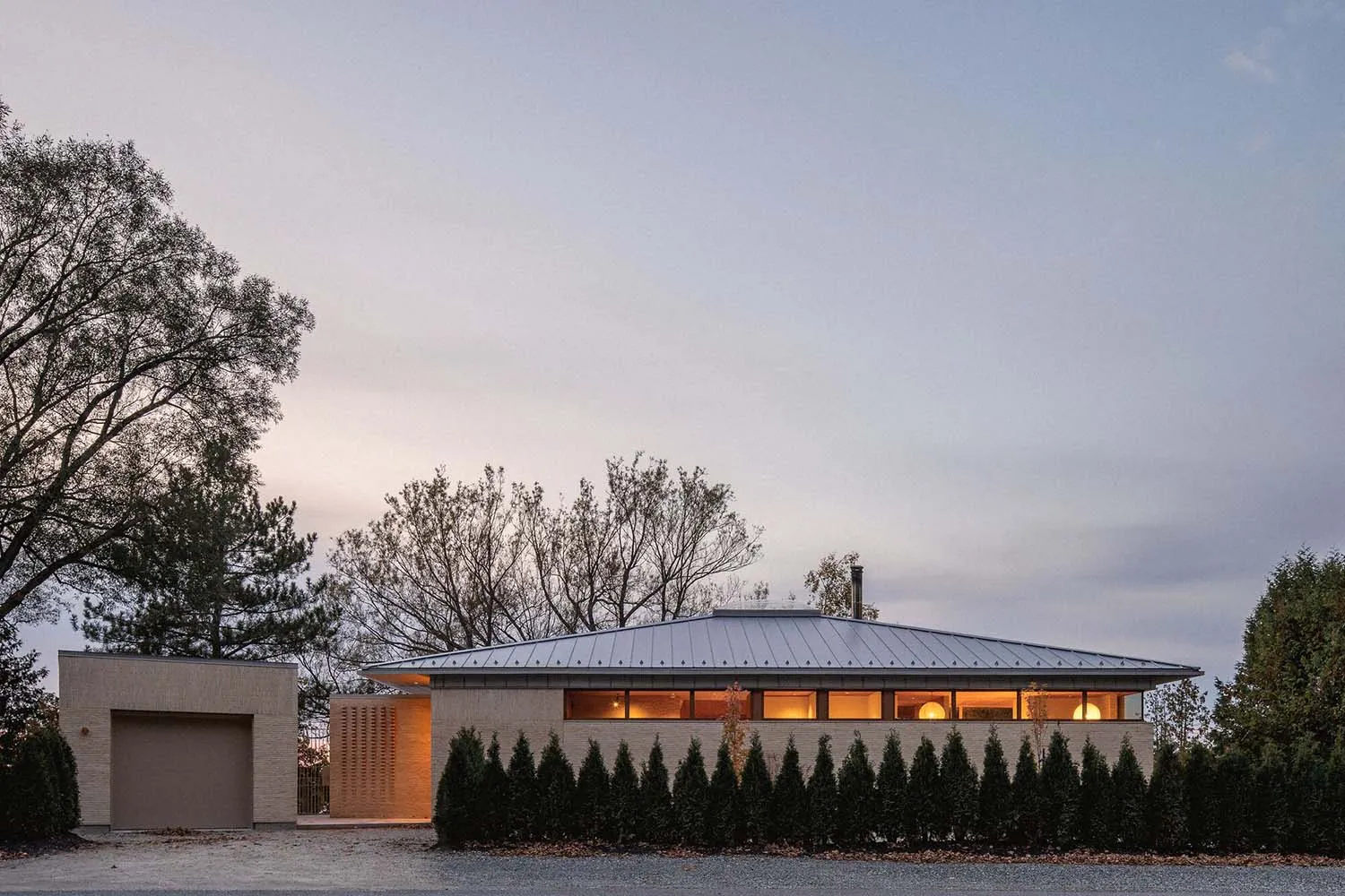 A modern residential house with a metal-clad exterior at dusk, featuring large windows with warm indoor lighting.