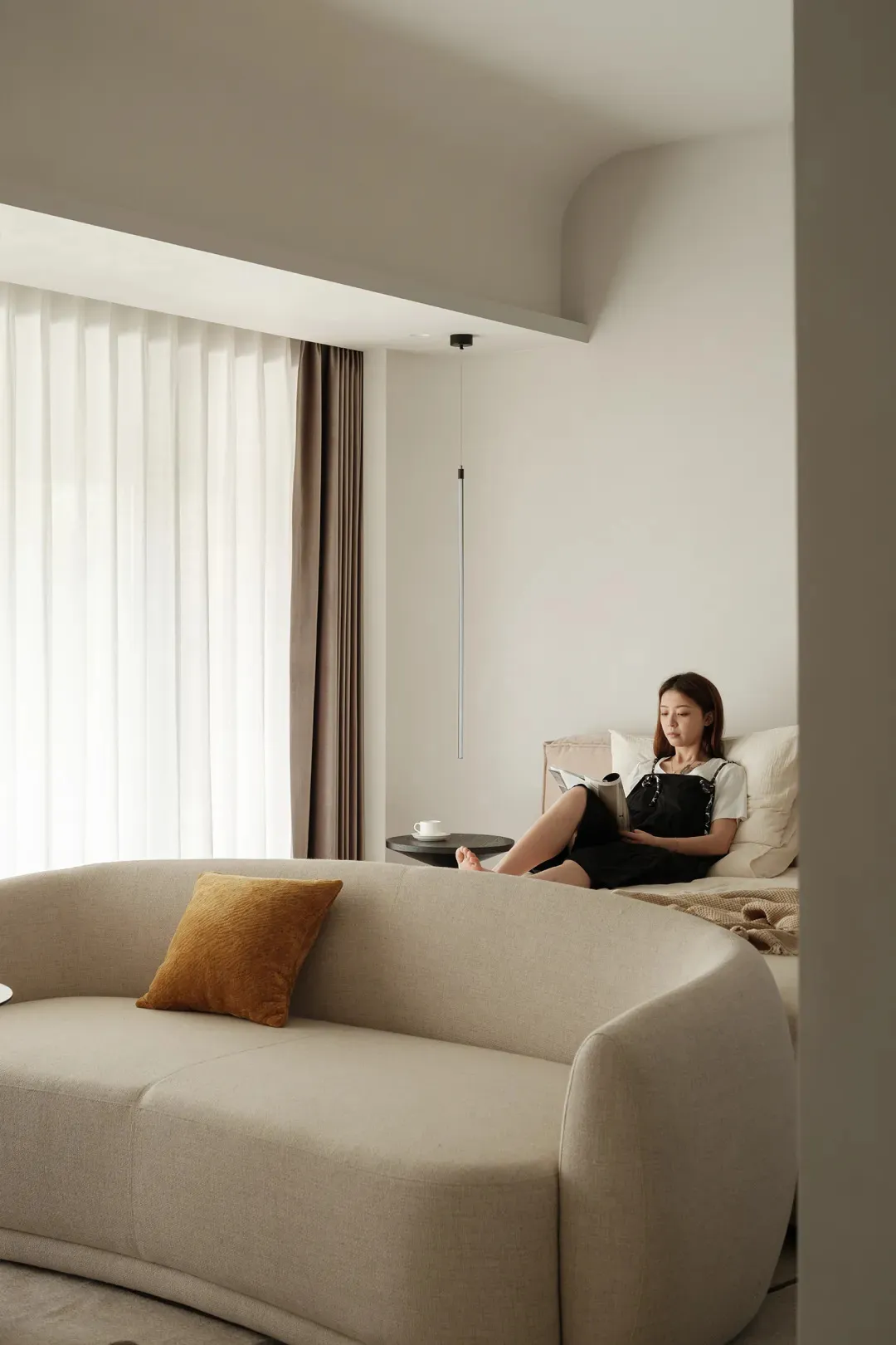 A minimalist living room with neutral-colored fabrics and a woman sitting in a bed reading a book.