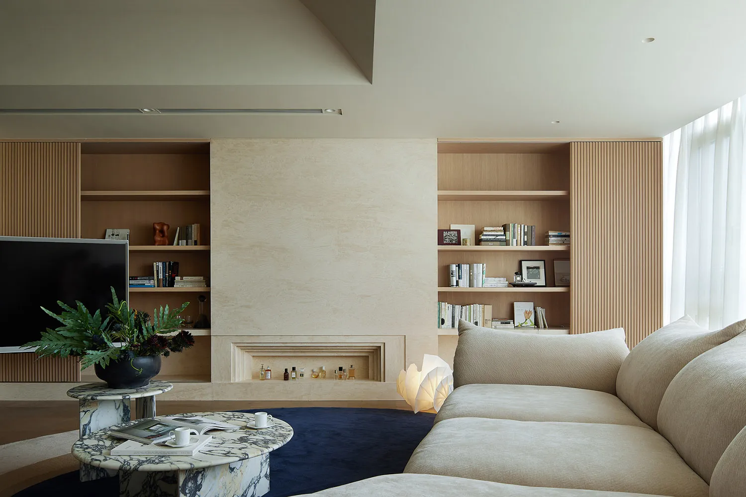 A modern minimalist living room with a beige sofa, marble coffee table, and wooden shelving.