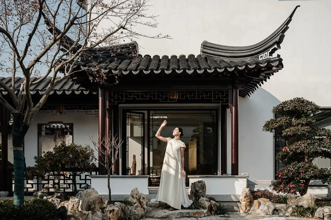 一位身穿白色洋裝的女子，站在傳統中式亭台前，亭台旁有假山庭園與盆栽樹木。