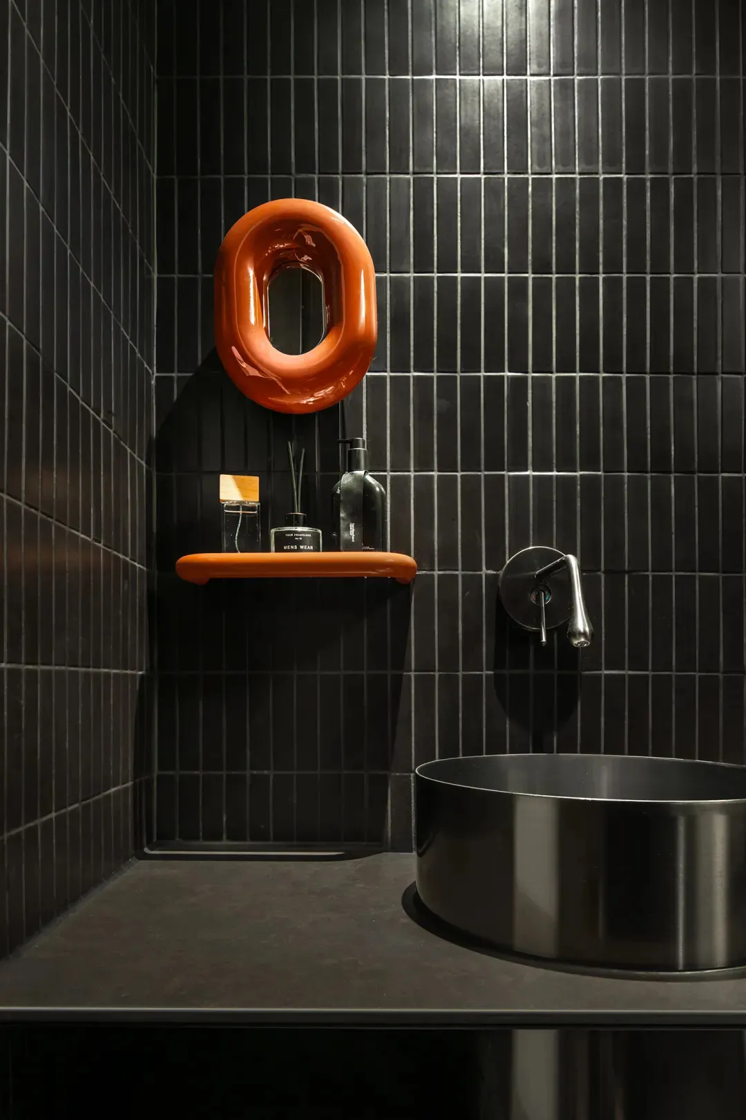Modern minimalist bathroom featuring dark tile walls, a metal sink, and a sleek shelf with bathroom essentials.