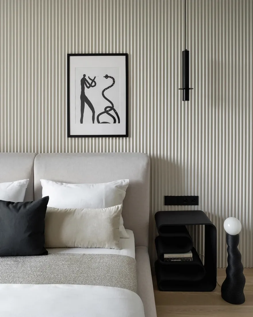 A modern minimalist bedroom with a light-colored upholstered bed, white and black pillows, and a geometric light fixture.