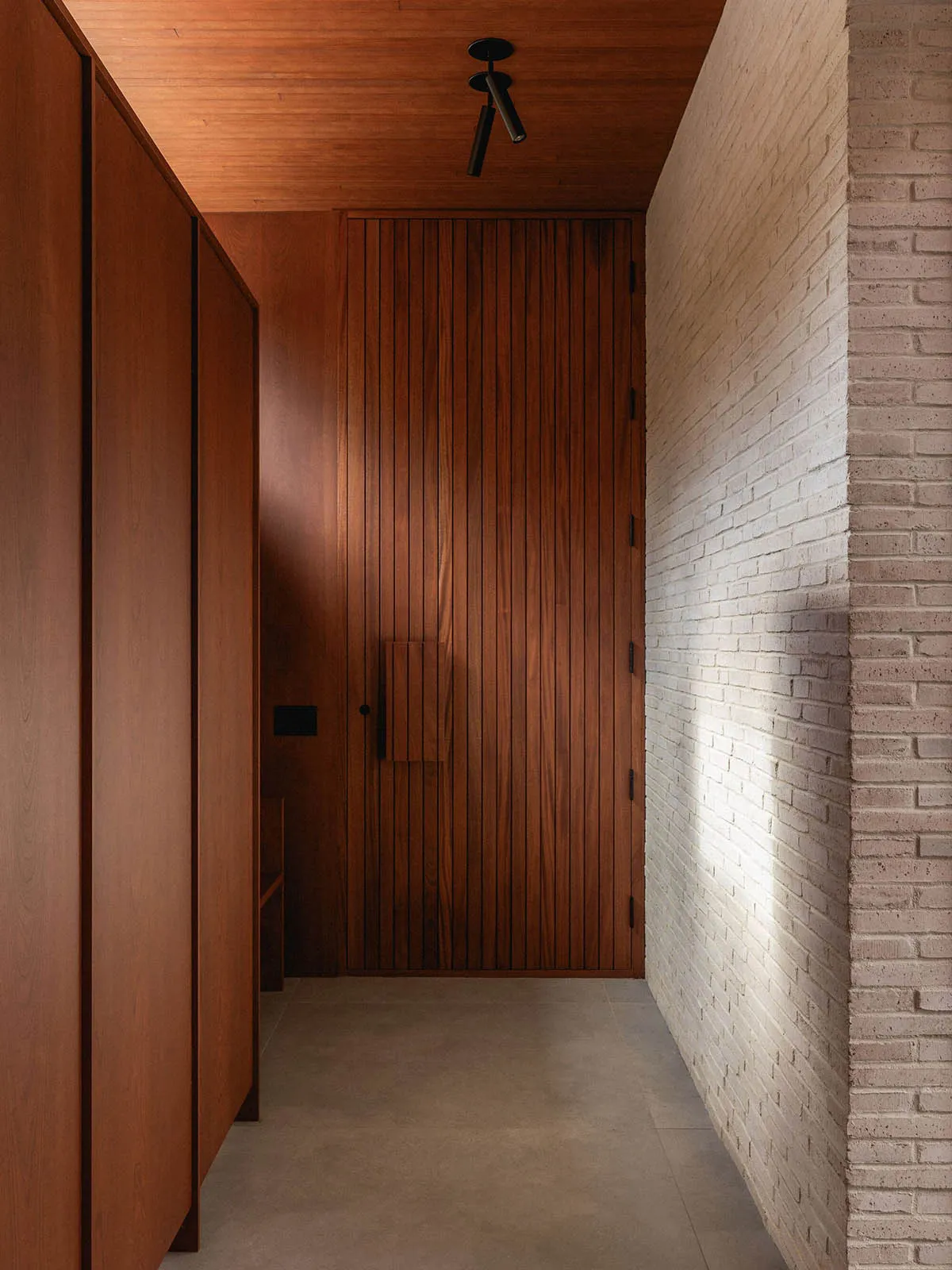 A modern minimalist entrance hall featuring a wooden door, concrete floor, and brick wall.