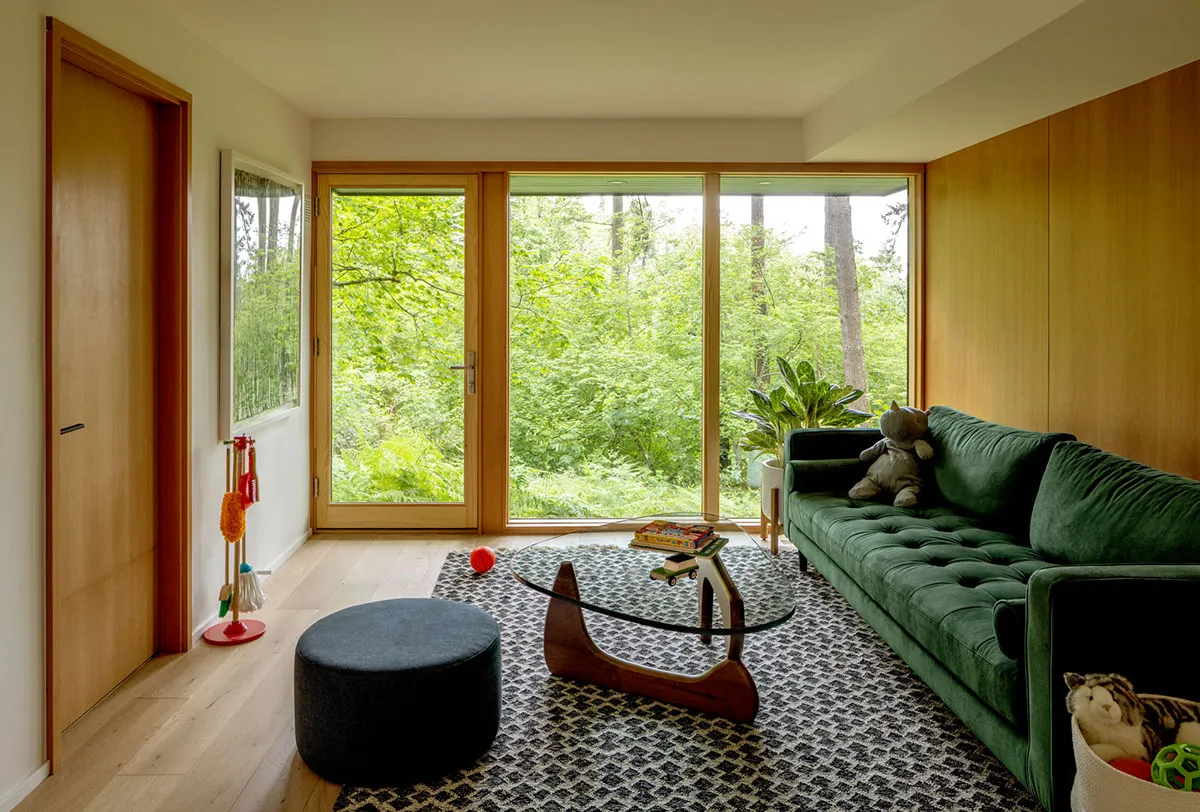 A modern minimalist living room featuring a green sofa, glass coffee table, and wooden flooring with a view of greenery outsi