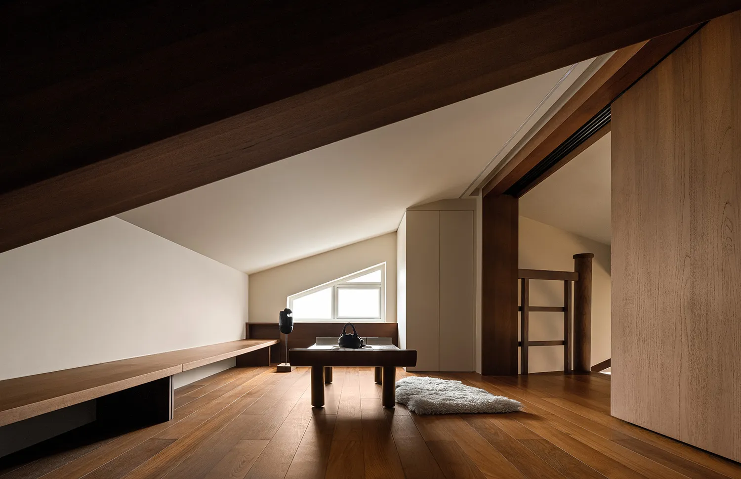 A minimalist living space with a wood-finished floor, featuring a bench, a table with a lamp, and a rug.