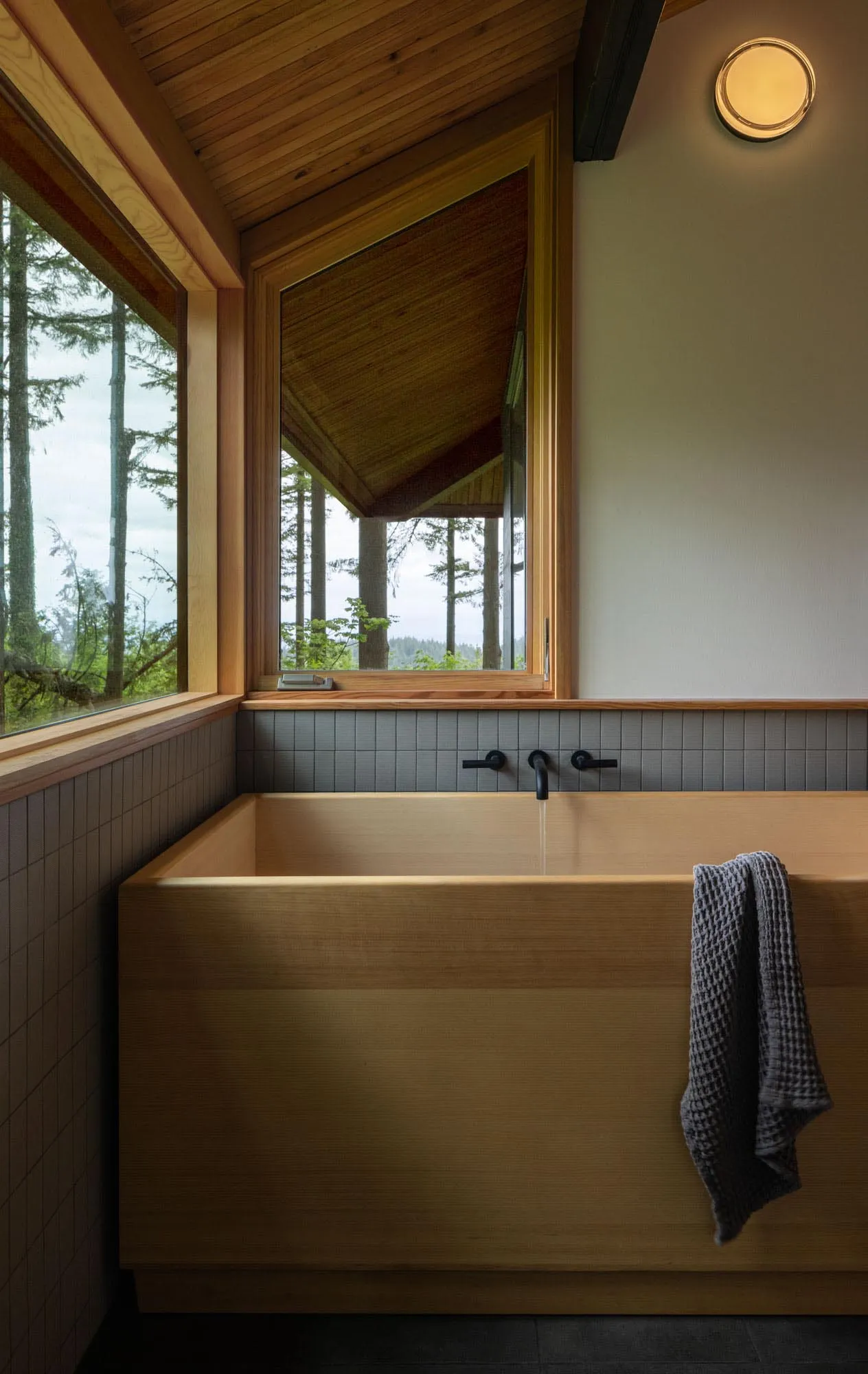 A modern minimalist bathroom with a wooden soaking tub and a view of the forest through a large window.