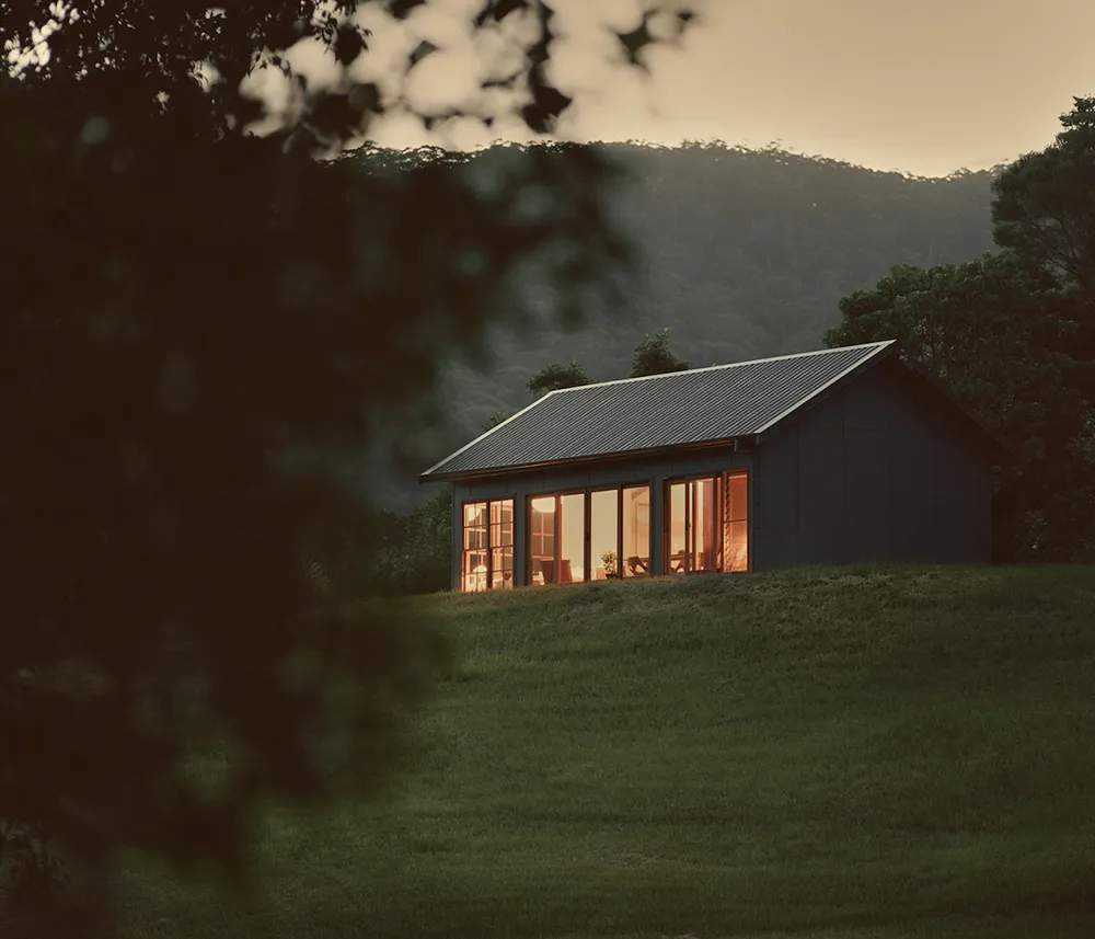 A minimalist house with metal and glass features, set against a scenic backdrop of trees and mountains.